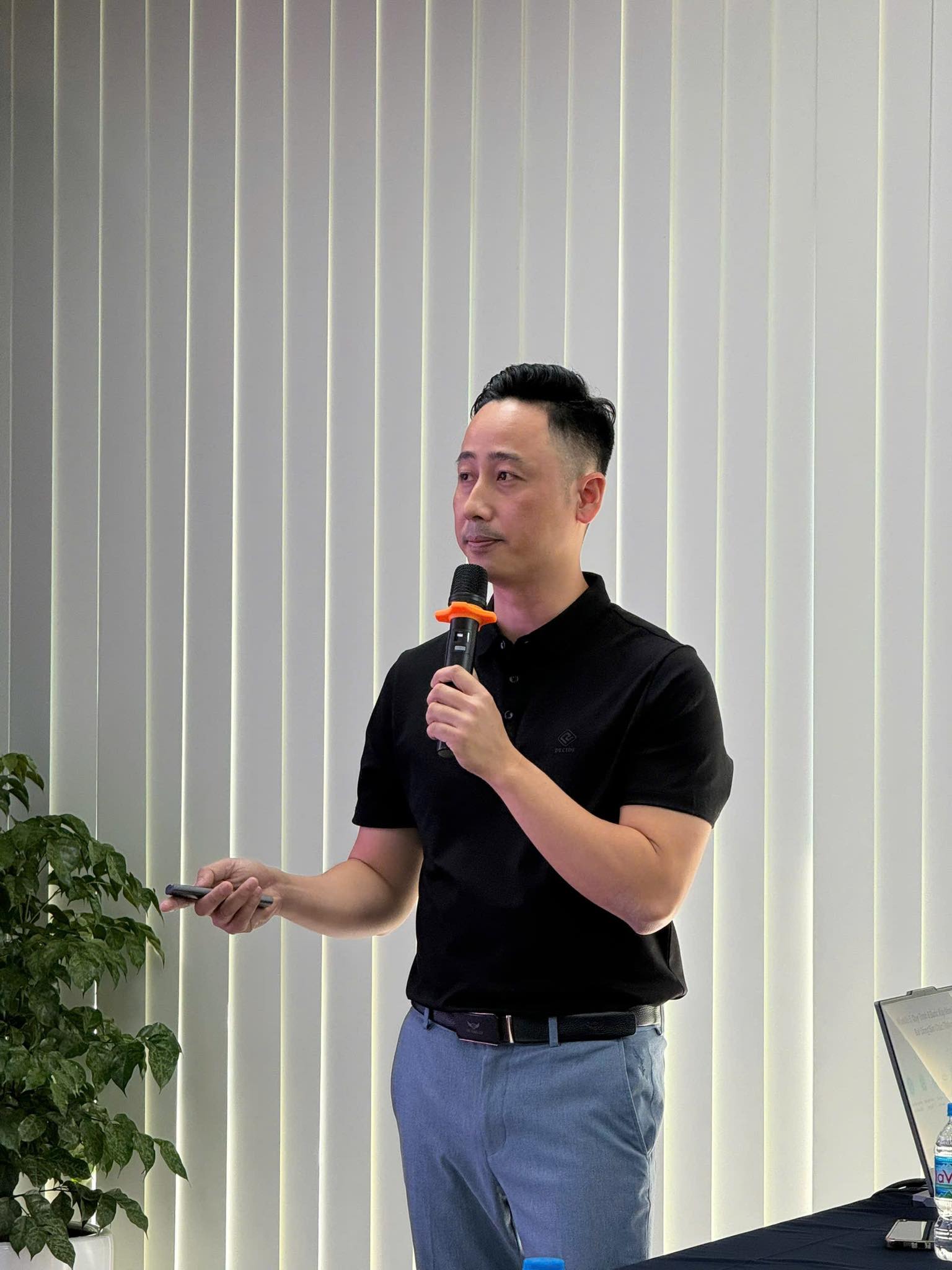 Đăng Dương mặc áo polo đen cầm micro và bút trình chiếu đứng chia sẻ trong một buổi đào tạo, bên cạnh có cây xanh và bàn đặt laptop