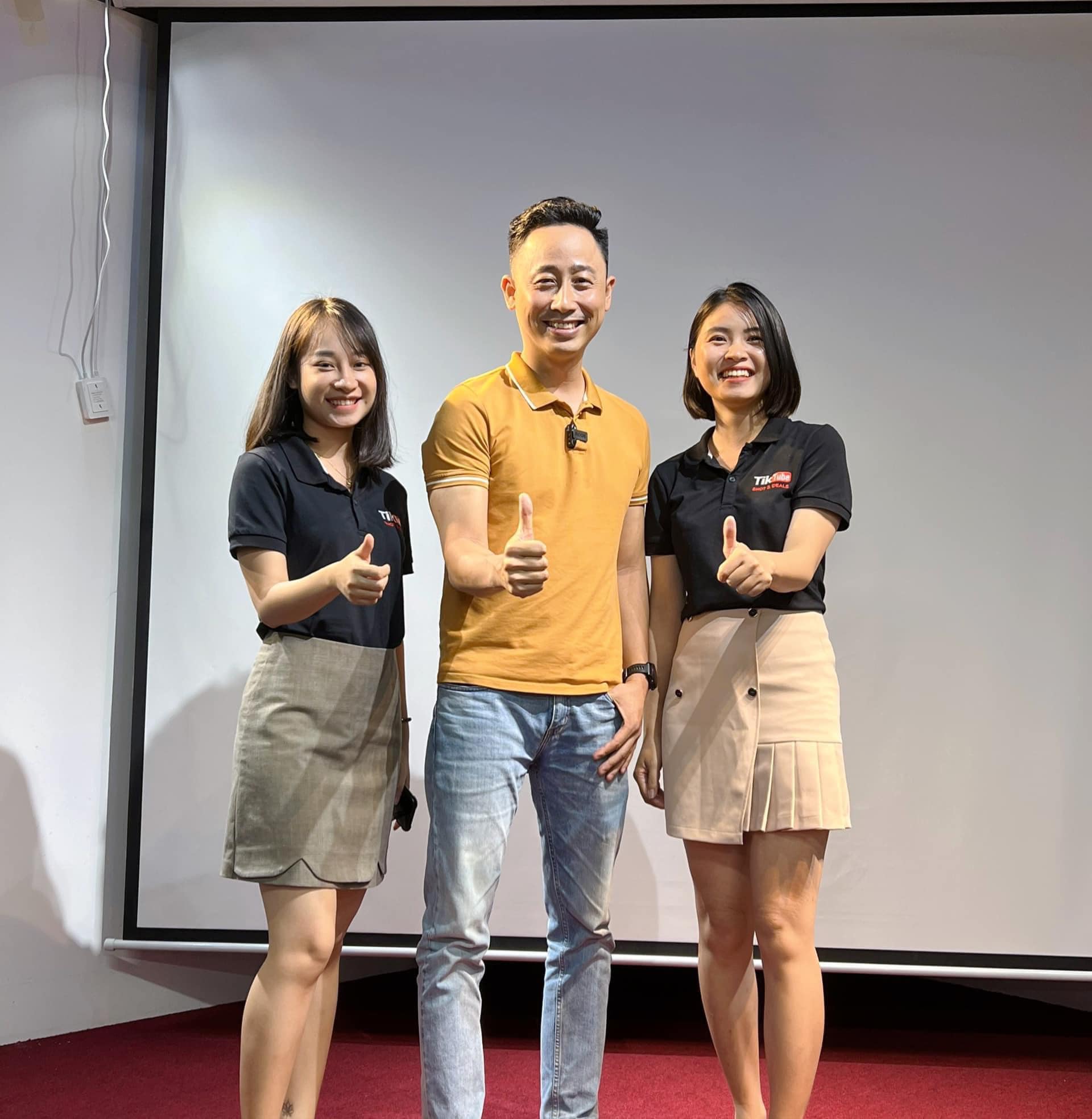 Đăng Dương mặc áo polo vàng đứng giữa hai đồng nghiệp nữ mặc áo polo đen, cả ba cùng giơ ngón tay cái trước phông nền màn chiếu