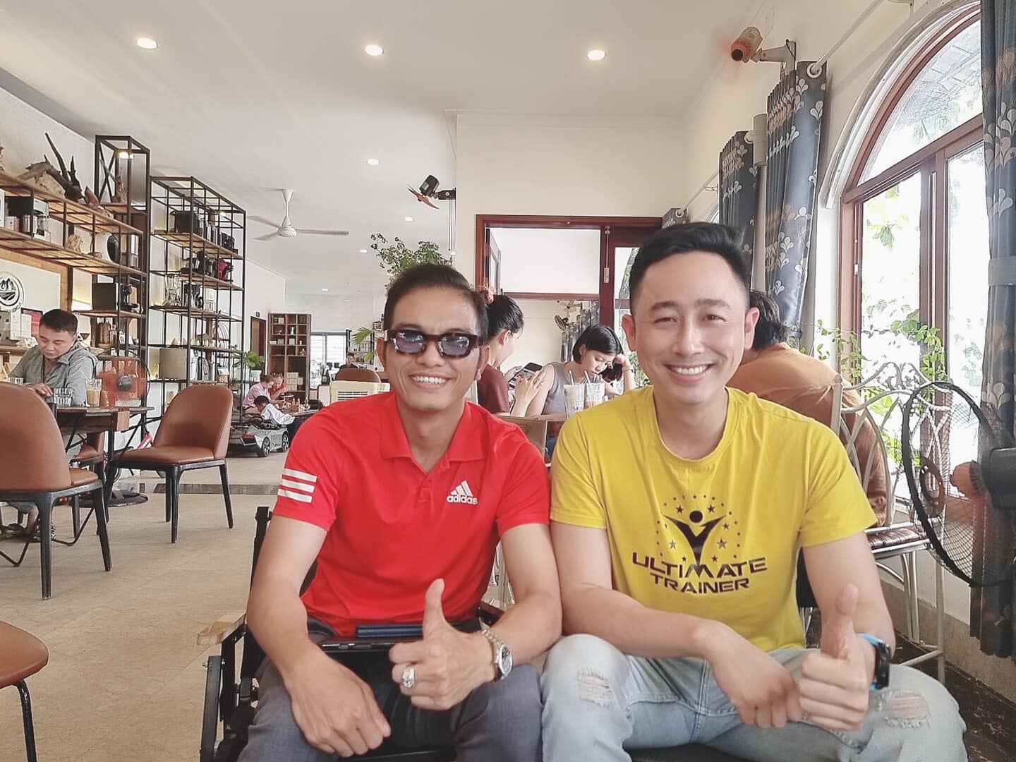 Đăng Dương mặc áo phông vàng ngồi chụp ảnh cùng một người đàn ông mặc áo polo đỏ đeo kính râm trong một quán cà phê, cả hai cùng giơ ngón tay cái