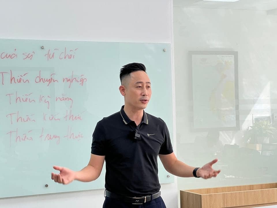 Đăng Dương mặc áo polo đen đứng giảng, hai tay xòe rộng, phía sau là tấm bảng kính ghi tay nhiều dòng chữ về nghề môi giới