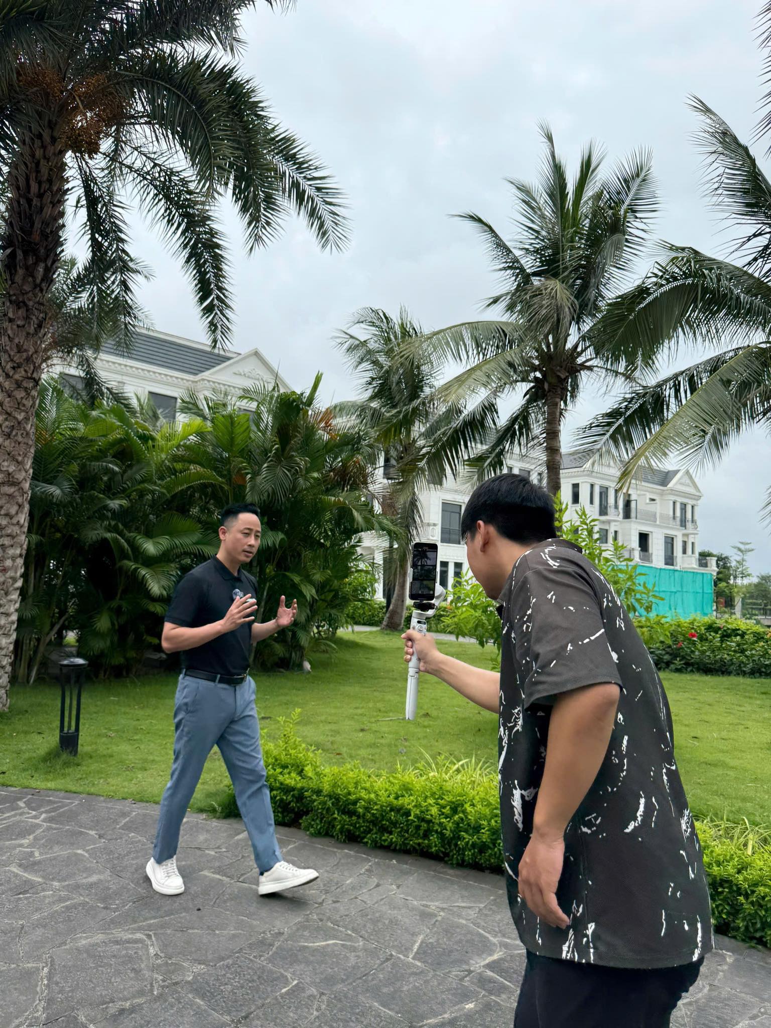 Đăng Dương mặc áo polo đen đứng nói trong sân vườn biệt thự nhiều cây dừa khi một người cầm điện thoại gắn tay cầm chống rung quay lại