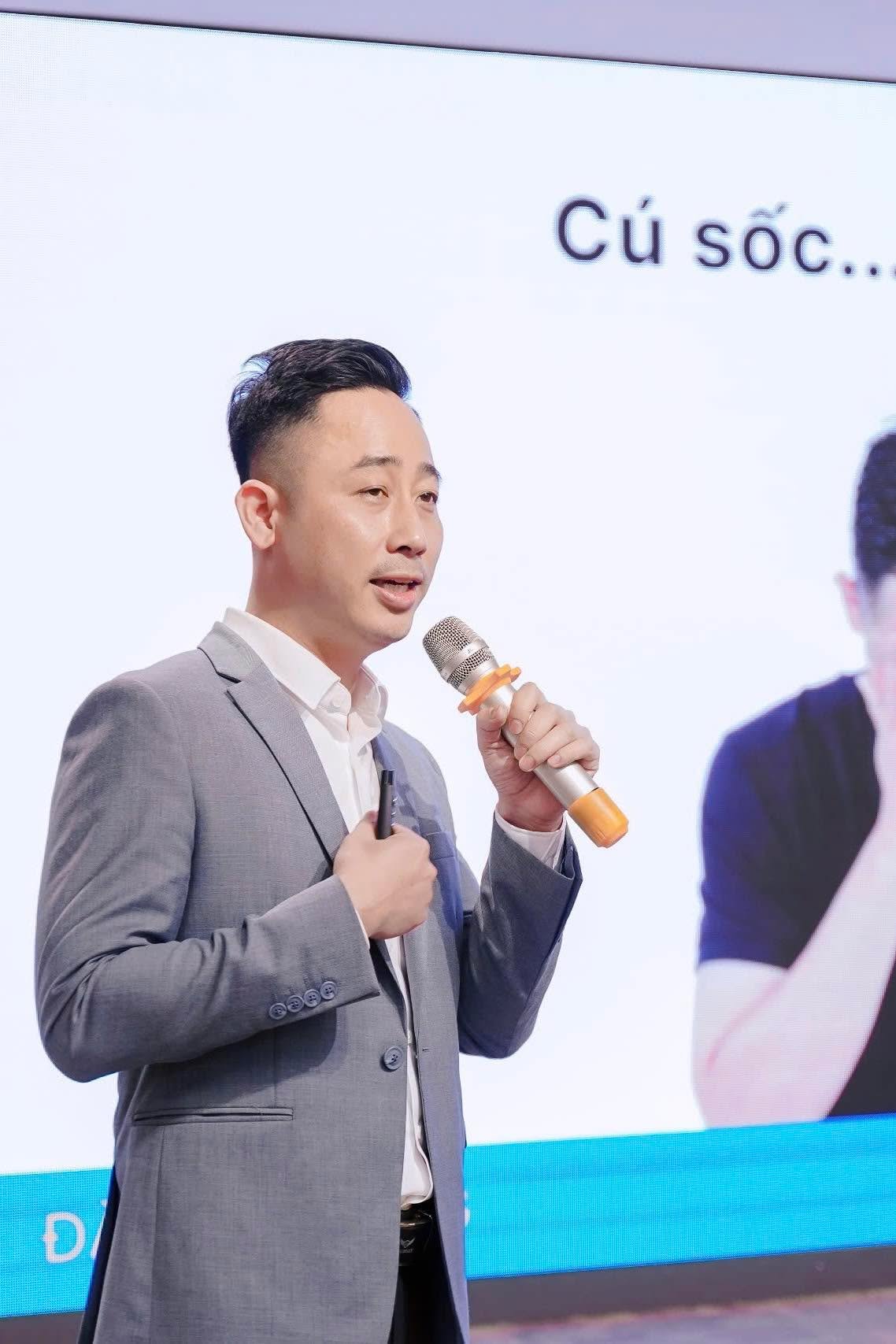 Đăng Dương mặc áo vest xám cầm micro nói trước màn hình chiếu trong một buổi chia sẻ