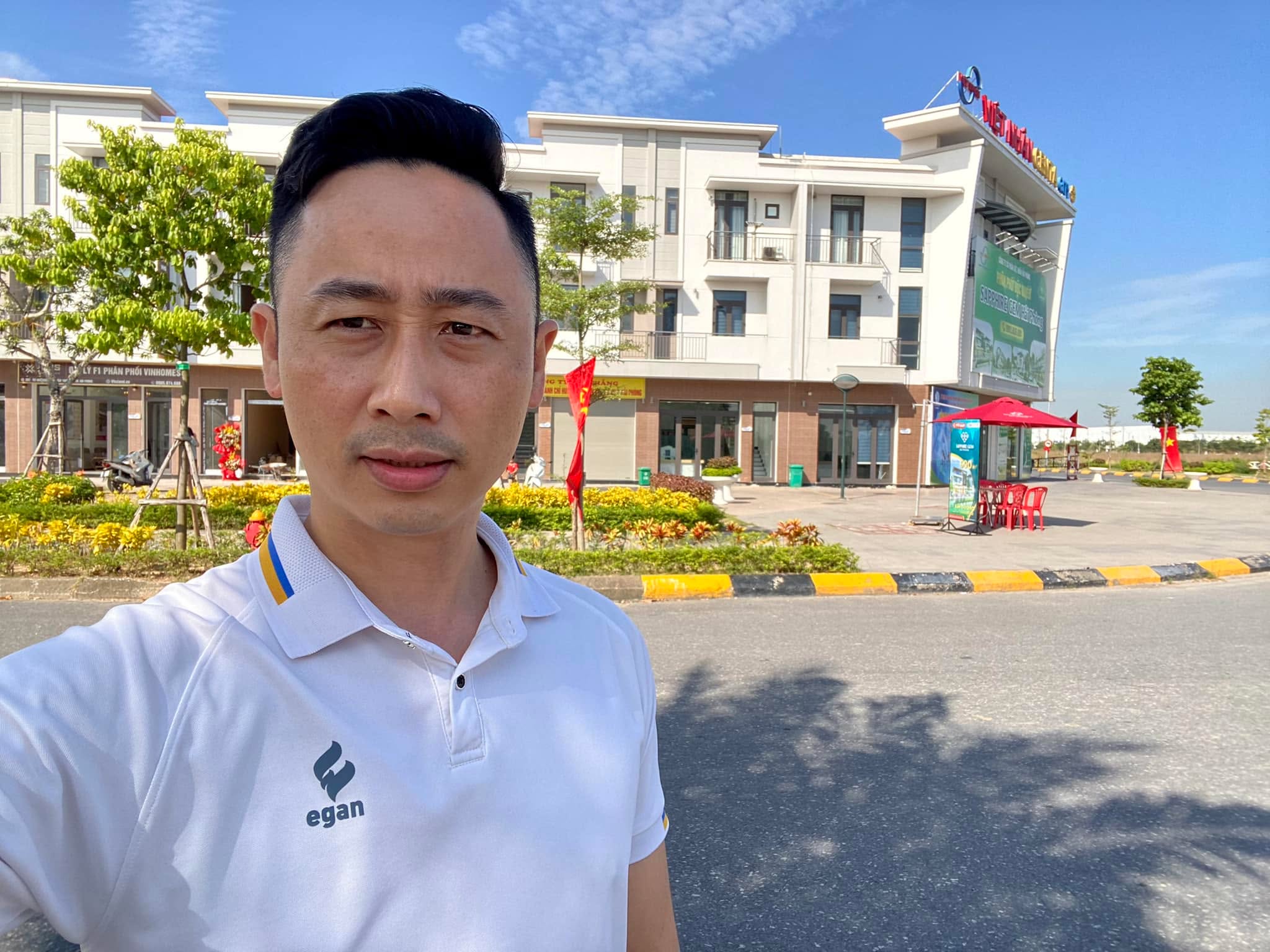 Đăng Dương mặc áo polo trắng đứng trước dãy nhà phố thương mại và một văn phòng giao dịch bất động sản trong khu đô thị