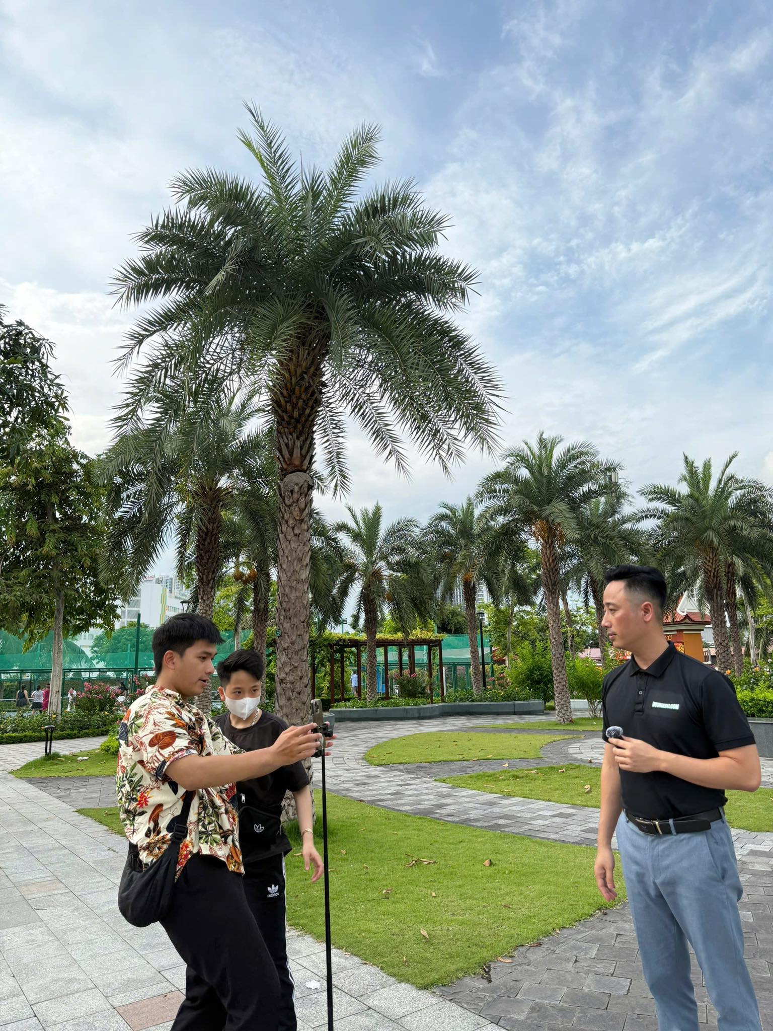 Đăng Dương mặc áo polo đen gắn micro cài áo, đứng cùng hai người hỗ trợ đang dựng điện thoại trên chân máy để quay video trong công viên