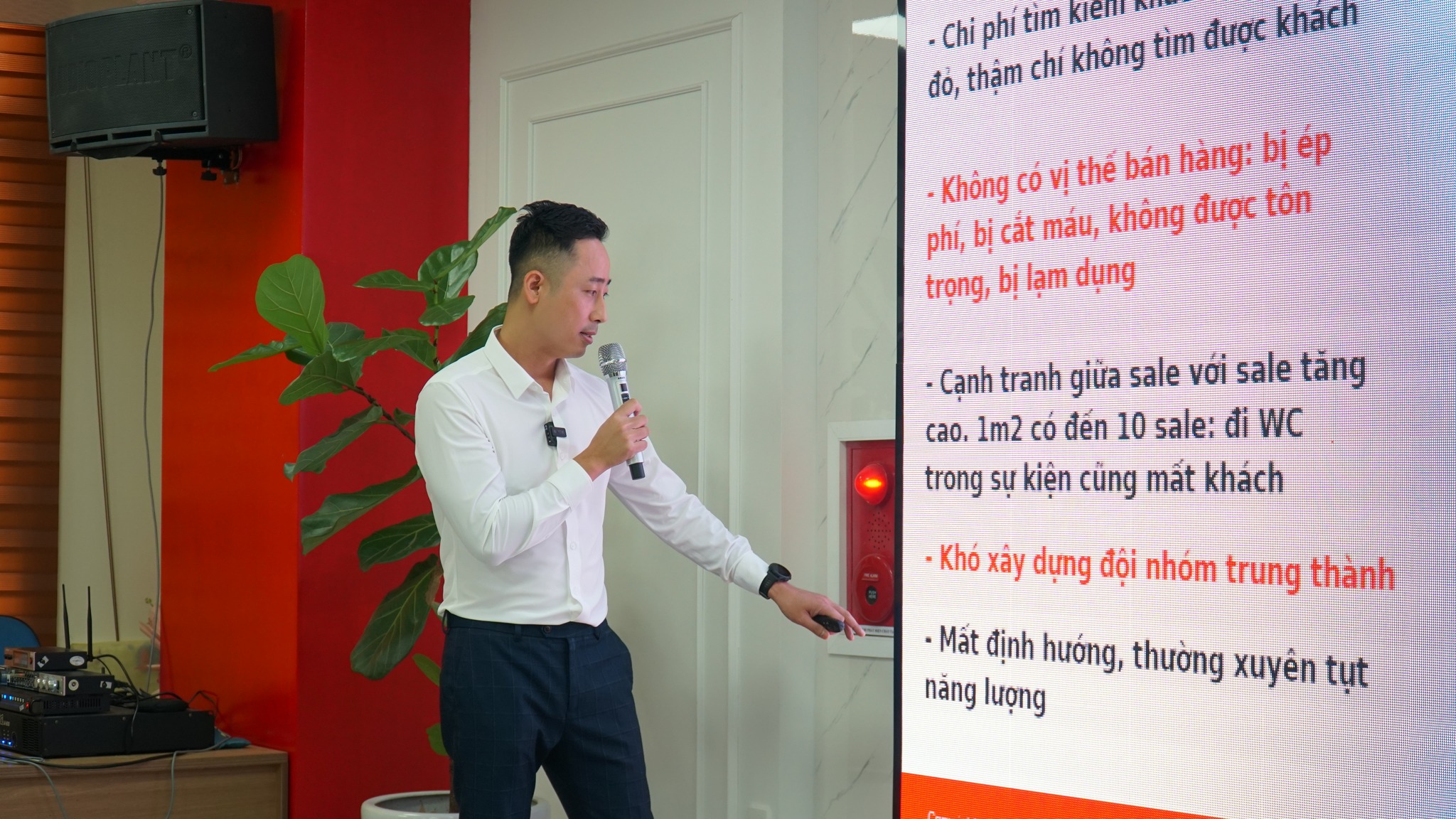 Đăng Dương mặc áo sơ mi trắng cầm micro, tay chỉ về màn hình trình chiếu lớn đang hiển thị các khó khăn của nghề sale bất động sản
