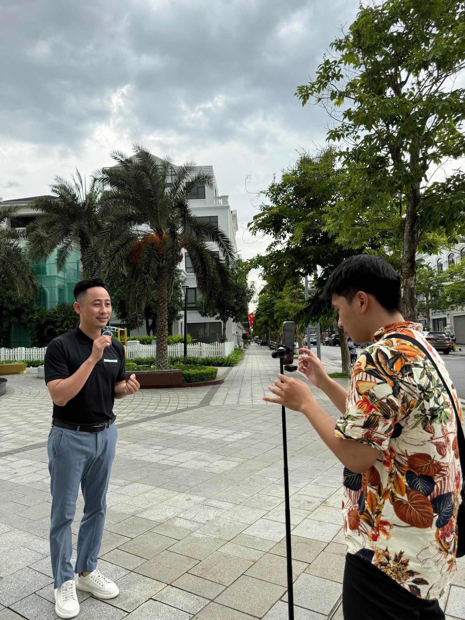 Đăng Dương mặc áo polo đen cầm micro đứng nói trên vỉa hè trong khu đô thị, phía trước một người mặc áo họa tiết đang cầm điện thoại gắn trên chân máy quay lại