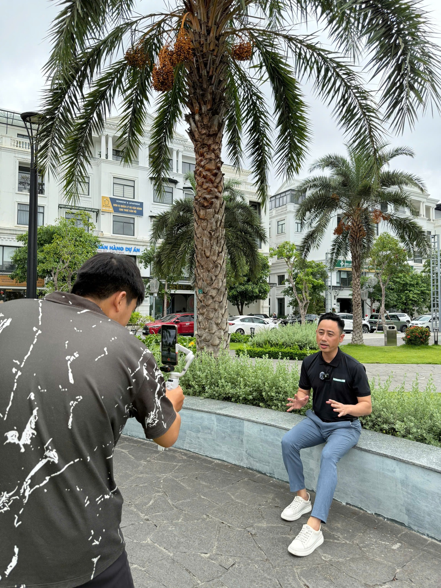Đăng Dương mặc áo polo đen ngồi trên bờ bồn cây dưới hàng cọ trong khu đô thị, nói trước một chiếc điện thoại gắn gimbal do một người đứng quay lại