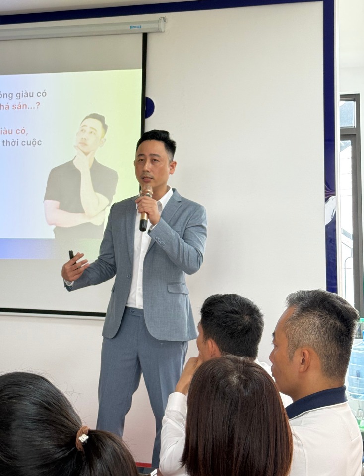 Đăng Dương mặc vest xám cầm micro đứng thuyết trình trước màn chiếu, phía dưới là các học viên ngồi nghe