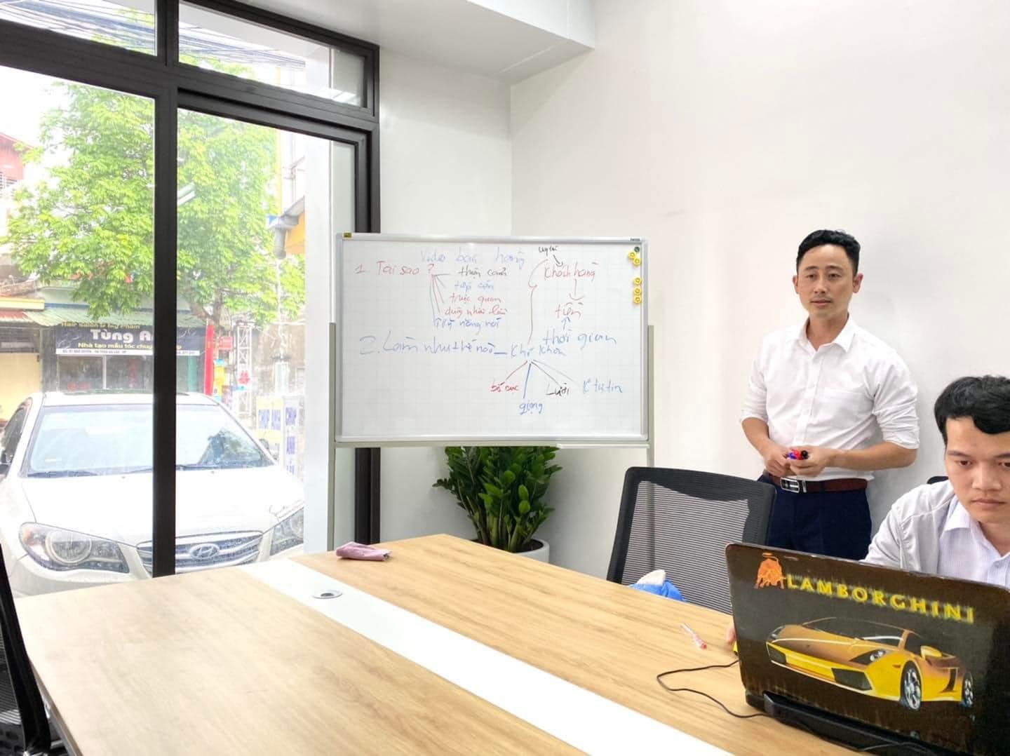 Đăng Dương mặc áo sơ mi trắng đứng cạnh tấm bảng trắng ghi sơ đồ về video bán hàng trong phòng làm việc, một người ngồi dùng laptop bên cạnh